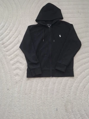 Svart hoodie från Polo Ralph Lauren - Säljer en svart hoodie från Polo Ralph Lauren med dragkedja, huva med dragsko och fickor framtill. Klassisk vit logga broderad på bröstet. Tillverkad i mjukt bomullsmaterial som är skönt att ha på sig. Perfekt för en avslappnad och stilren look.