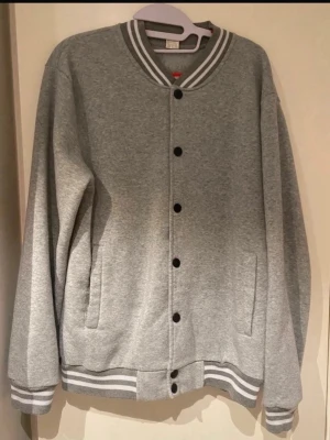 Grå collegehoodie med knappar - Snygg grå hoodie i collegestil med svarta knappar framtill och vita/grå ränder på krage, ärmslut och nederkant. Tillverkad i 95% polyester och 5% elastan för en mjuk och stretchig känsla. Två fickor framtill och klassisk baseboll-look. Herrmodell S. Är nopprig men det kan man noppra bort. 