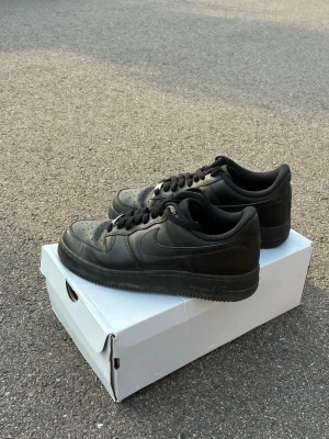 Svarta Nike Air Force 1 sneakers - Säljer ett par klassiska svarta Nike Air Force 1 sneakers med låg profil och perforerad tå. Skorna har en robust gummisula, svart läderöverdel och ikonisk Swoosh på sidan. Perfekta för dig som vill ha en clean och tidlös look.