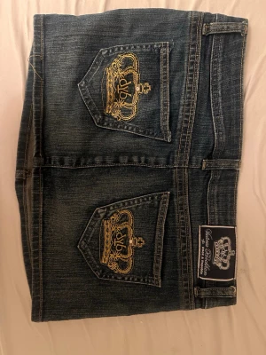 Mörk jeanskjol med guldbroderi - Snygg mörkblå jeanskjol från Victoria Beckham for rock & republic med två bakfickor prydda av guldbroderade kronor. Klassisk femficksmodell med knapp och dragkedja framtill. Perfekt för dig som gillar streetwear och detaljer som sticker ut.