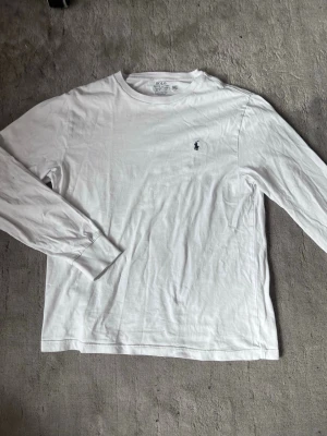 Vit långärmad tröja Polo Ralph Lauren - Vit långärmad tröja från Polo Ralph Lauren med rund hals och mörkblå broderad logga på bröstet. Tillverkad i mjuk bomull för en clean och enkel look. Perfekt att styla med jeans eller linnebyxor