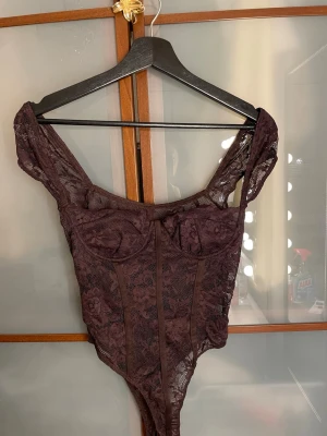 Brun spetsbody med breda axelband - Snygg brun body i genomskinlig spets med blommönster och breda axelband. Bodyn har bygelkupor och markerade sömmar framtill för en extra formande effekt. Perfekt för dig som vill ha något både trendigt och bekvämt.
