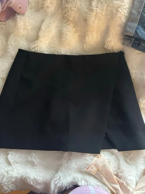 Svart skort med omlottlook - Stilren svart skort med omlottkänsla framtill. Klassisk rak passform och mjukt, vävt tyg som ger en snygg siluett. Perfekt för dig som vill ha kjol-looken men med shortskomfort. Enkel att matcha med olika toppar och accessoarer.