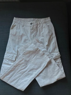 Beiga cargobyxor med fickor - Snygga beiga cargobyxor med flera praktiska fickor på sidorna och bak. Byxorna har raka ben och klassisk cargomodell med knapp och dragkedja framtill. Tillverkade i ett mjukt bomullstyg som är bekvämt att bära hela dagen.