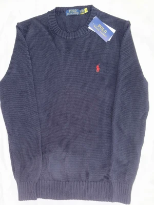 Mörkblå stickad tröja Polo Ralph Lauren - Klassisk mörkblå stickad tröja från Polo Ralph Lauren med rund halsringning och röd broderad logga på bröstet. Tröjan har ribbade muddar vid ärmslut och nederkant, vilket ger en snygg och tidlös look. Perfekt för lager på lager under kalla dagar.