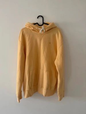 Hollister hoodie gul - Snygg gul hoodie med stor huva och vita dragsnören. Klassisk känguruficka framtill och avslappnad passform. Perfekt för chill dagar eller när du vill ha en enkel men stilren look. Tillverkad i mjukt material för extra komfort.