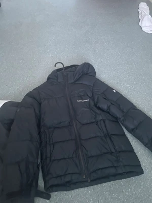 Svart dunjacka med huva - Säljer en svart dunjacka från Helly Hansen med huva och dragkedja framtill. Jackan har quiltad design och logga på bröstet samt ärmen. Perfekt för kalla vinterdagar och riktigt skön att bära.