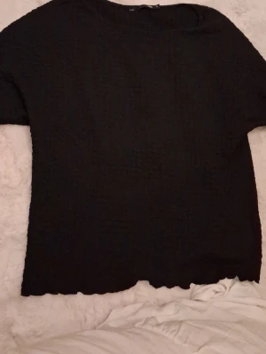 Svart strukturerad topp - En svart topp med strukturerat tyg och rund hals. Toppen har korta ärmar och en enkel, stilren design som passar till många olika looks. Perfekt för dig som gillar minimalistisk stil.