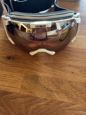 Bliz skidglasögon med extra lins - Säljer ett par vita skidglasögon från Bliz med spegelreflekterande lins och extra gul lins för olika ljusförhållanden. Skön passform med bred resår och mjuk skumkant. Perfekt för dig som vill ha stilrena och funktionella goggles i backen.
