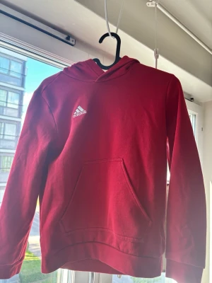 Röd hoodie från Adidas med huva - Snygg röd hoodie från Adidas med klassisk vit logga på bröstet. Hoodien har en stor känguruficka framtill och en skön huva. Perfekt för en avslappnad och sportig stil. Tillverkad i mjukt material som känns bekvämt hela dagen. OBS BARN TRÖJA 140CM!
