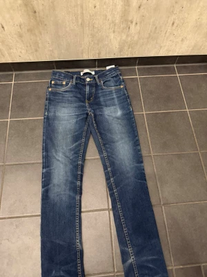 Blå raka jeans - Säljer ett par klassiska blå jeans med raka ben och snygga slitningar framtill. Jeansen har fem fickor, kontrastsömmar och en liten röd etikett på bakfickan. Tillverkade i denim med en cool tvättad look.