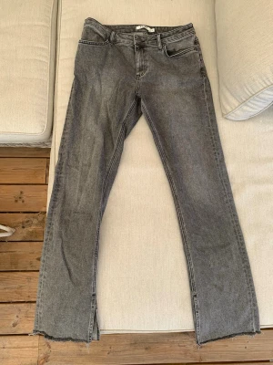 Grå bootcut jeans med slit kant - Säljer ett par grå jeans med bootcut passform med slit nertill och fransig kant. Jeansen har klassisk femficksmodell och låg midja. Materialet är denim i bomull och färgen är tvättad grå. Perfekta för en avslappnad och trendig look.