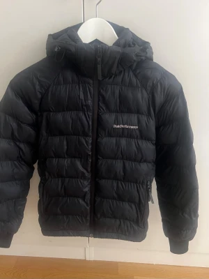 Svart dunjacka Peak Performance - Snygg svart dunjacka från Peak Performance med huva och dragkedja framtill. Jackan har två sidofickor med dragkedja och diskret logga på bröstet. Perfekt för kalla vinterdagar tack vare det fluffiga dunfodret och den klassiska quiltade designen. Den har en lagning på framsidan, se bild