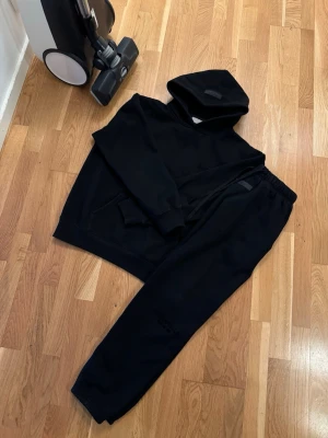 Essentials tracksuit - Säljer ett svart loungewear-set med hoodie och matchande byxor. Hoodien har huva och magficka, byxorna har resår i midjan och vid benslut. Perfekt för chill dagar hemma eller när du vill vara bekväm. Materialet känns mjukt och skönt mot huden.