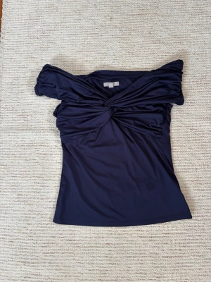 Marinblå offshoulder topp H&M - Säljer en marinblå offshoulder topp från H&M i mjukt och stretchigt material. Toppen har snygg twistad detalj framtill och sitter tight över kroppen. Perfekt för dig som vill ha en trendig och stilren look.