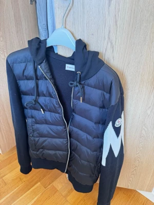 Svart pufferjacka med huva från Moncler. Tar även byten - Säljer en svart pufferjacka från Moncler med stickade ärmar och huva. Jackan har dragkedja framtill, fickor och ett stort vitt mönster på ena ärmen. Perfekt för kalla dagar och snygg att matcha med jeans. Med qr kod och tagg i armen 