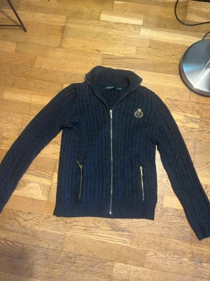 Mörkblå kabelstickad tröja Lauren - Mörkblå kabelstickad tröja med dragkedja framtill och två fickor med guldfärgade dragkedjor. Tröjan har hög krage och ett broderat RL-emblem i guld på bröstet. Perfekt för dig som gillar klassisk och stilren look. Storleken är M men passar även S. Notera att lappen sitter löst. 