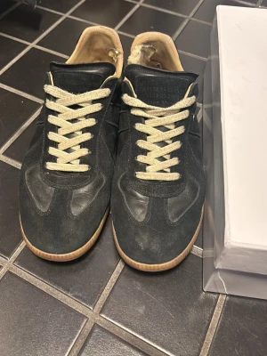 Maison margielas - Säljer ett par svarta sneakers med beige sula och vita snören. Skorna har en klassisk låg siluett och är tillverkade i mocka och läder. Insidan är ljus och de har en stilren look som passar till det mesta.