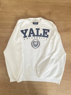 Vit sweatshirt med Yale-tryck - Vit sweatshirt med stort blått YALE UNIVERSITY-tryck och emblem på bröstet. Tröjan har rund halsringning och långa ärmar. Perfekt för en avslappnad och klassisk college-look.