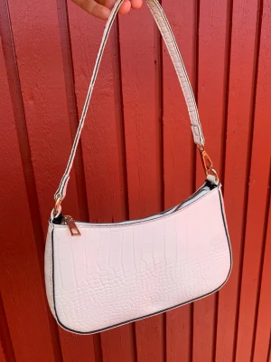 Vit axelväska i crocopräglat material - Snygg vit handväska med axelrem och crocopräglat mönster. Väskan har guldfärgade detaljer och dragkedja upptill. Perfekt storlek för det viktigaste och ger en trendig touch till din outfit.
