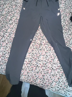 Mörkgrå mjukisbyxor med dragsko - Snygga mörkgrå mjukisbyxor med smal passform och dragsko i midjan. Byxorna har fickor med dragkedja och reflekterande detaljer. Perfekta för chill eller träning, tillverkade i ett mjukt och stretchigt material.