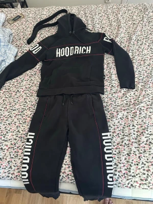 Svart Hoodrich set med tryck - Säljer ett svart Hoodrich set med hoodie och byxor. Setet har vita tryck på bröstet och benen samt röda detaljer längs sömmarna. Hoodien har huva med snörning och magficka, byxorna har resår och dragsko i midjan. Perfekt för en avslappnad streetwear-look.
