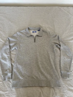 Grå halvzip tröja med krage - En stilren grå sweatshirt med halv dragkedja och klassisk krage. Tröjan har långa ärmar, ribbade muddar och är gjord i mjukt sweatshirtmaterial. Perfekt för en avslappnad och snygg look.