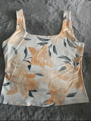 Blommig linnetopp i polyester - Säljer en söt blommig linnetopp med stora ljusgula blommor och gröna blad på vit botten. Toppen har breda axelband och fyrkantig ringning. Tillverkad i mjuk polyester med lite stretch för skön passform. Storlek L men är mer som M.