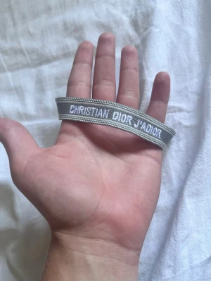 Grått Christian Dior J'Adior armband - Snyggt grått tygarmband från Christian Dior med texten 'J'ADIOR' broderad i vitt. Armbandet har en enkel och stilren design med justerbar knytning och tofsar i ändarna. Perfekt accessoar för att lyfta din outfit.