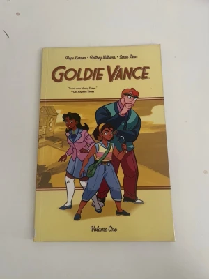 Goldie Vance Volume One - Goldie Vance Volume One är en grafisk roman med färgglad tecknad framsida som visar tre karaktärer i äventyrsstil. Omslaget är gult och boken är i pocketformat. Perfekt för dig som gillar mysterier och ungdomsserier.