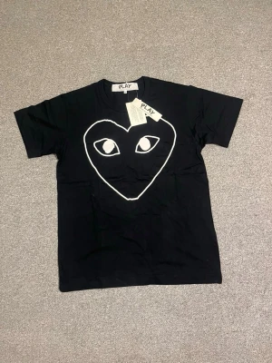 Svart Comme des Garçons Play t-shirt - Svart t-shirt från Comme des Garçons Play med stort vitt hjärta och ögon på bröstet. Klassisk rund hals och korta ärmar. Tillverkad i mjuk bomull och har en clean, stilren look med tydlig streetwear-vibe.