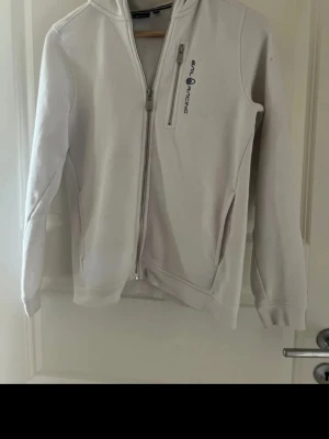 Vit hoodie från Sail Racing - Snygg vit hoodie från Sail Racing med dragkedja framtill och två fickor. Har en liten logga på bröstet och en huva. Tillverkad i mjukt material som känns skönt mot huden. Perfekt för en avslappnad och sportig stil. Ny pris 799 ✅ 