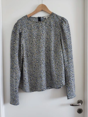 Blommig långärmad blus i blått och vitt - Snygg blus med småblommigt mönster i blått, vitt och beige. Modellen har rund halsringning och långa ärmar med lätt puff vid axlarna. Tunn och luftig kvalitet, perfekt till jeans eller kjol. Stängs med knapp i nacken.