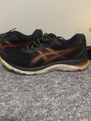 Svarta Asics sneakers  - Snygga svarta sneakers från Asics med orange logga och detaljer, samt gröna och svarta snören. Ovandelen är i mesh för bra ventilation och sulan är vit med inslag av gult och orange. Perfekta för dig som gillar sportig stil och komfort.