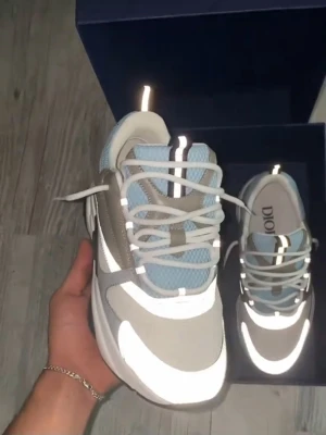 Dior sneakers i grått och blått - Snygga Dior sneakers i grått, vitt och ljusblått med reflekterande detaljer. Skorna har en chunky sula, meshpaneler och vita skosnören. Perfekt för dig som gillar exklusiva och trendiga sneakers med modern design.