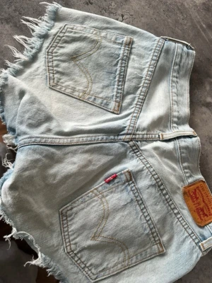 Levi’s 501 Original Jeansshorts – Ljus tvätt – S/M - Ljusblå Levi's 501 jeansshorts med hög midja och råa fransiga benslut. Klassisk femficksmodell i bomull med knappgylf och avslappnad loose fit. Perfekta för sommardagar och ger en chill vintagekänsla.