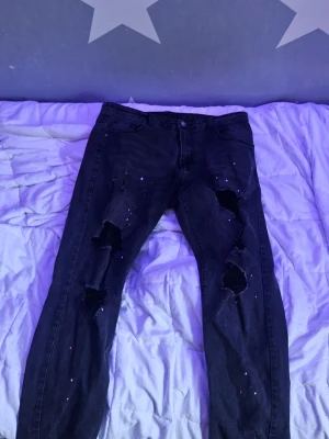 Svarta slitna jeans med färgstänk - Säljer ett par svarta jeans med stora slitningar på båda benen och små färgstänk i olika färger. Jeansen har en smal passform och klassisk femficksdesign. Perfekta för dig som gillar en edgy och trendig stil.