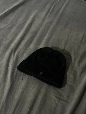  Ralph Lauren - Snygg svart mössa från Polo Ralph Lauren med ribbad kant och liten röd logga framtill. Klassisk och enkel design som passar till det mesta. Tillverkad i mjukt stickat material som håller dig varm under kalla dagar.