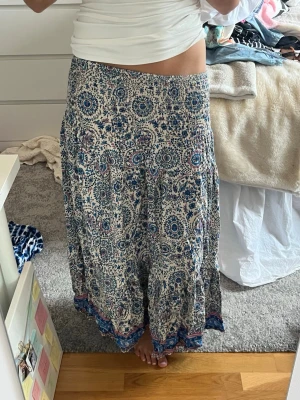 Blommig långkjol i blått och vitt - Lågmidjad långkjol med blommigt mönster i blått och vitt samt inslag av lila. Kjolen har en bred resår i midjan och ett flowy fall. Materialet känns lätt och luftigt, perfekt för varmare dagar. Fina blå detaljer längs nederkanten ger extra stil. Jag har storlek 36/s och den passar mig men den är väldigt stretchig så passar troligtvis 34-40 oxå