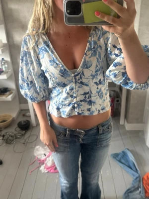 Blommig croppad blus med puffärm - Säljer en superfin croppad blus med blått blommönster på vit botten. Blusen har v-ringning, puffiga trekvartsärmar och knappar framtill. Perfekt för dig som vill ha en trendig och somrig look!