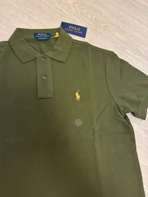 Olivgrön pikétröja från Polo Ralph Lauren - Snygg olivgrön pikétröja från Polo Ralph Lauren med klassisk krage och två knappar framtill. Tröjan har det ikoniska broderade logotypen i gult på bröstet och är tillverkad i mjuk bomull. Perfekt till jeans eller shorts för en clean look.