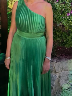 Grön plisserad one shoulder-klänning - Snygg långklänning i plisserat grönt tyg med one shoulder-design och markerad midja. Klänningen har ett elegant fall och passar perfekt när du vill sticka ut med en färgstark look.
