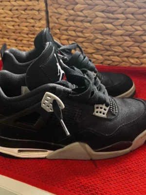 Svarta Jorden 4  - Säljer ett par svarta sneakers med vita och grå detaljer samt meshpaneler på sidorna. Skorna har en hög plös med Jumpman-logga och grova svarta skosnören. Sulan är vit och grå med synlig dämpning. Perfekta för dig som gillar streetwear och sportig stil.