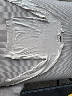 Beige stickad tröja  - En stilren beige stickad tröja med rund hals och långa ärmar. Tröjan har ribbade muddar vid ärmslut och nederkant, vilket ger en snygg och enkel look. Perfekt att matcha med jeans eller kjol.