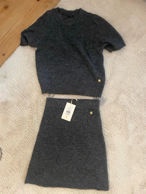 Mörkgrå stickat set - Mysig mörkgrå stickad topp med korta ärmar och rund hals, och stickad kjol till. Toppen har en ribbad kant nedtill och en liten guldfärgad knappdetalj vid nederkanten. Perfekt för dig som gillar en enkel men stilren look. Med prislappen kvar, aldrig använt!
