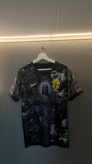 Svart Brasil Nike fotbollströja - Snygg svart fotbollströja från Nike med Brasilien-tema. Tröjan har ett detaljerat grafiskt tryck med Kristusstatyn och brasilianska symboler, samt gult Nike-märke och Brasil-logga med broderade stjärnor. Perfekt för dig som älskar fotboll och vill sticka ut på planen.