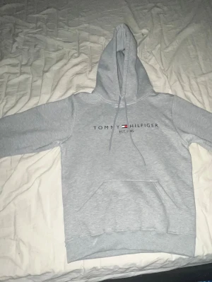 Ljusgrå hoodie med tryck - Ljusgrå hoodie med stor magficka och huva med dragsnören. Tryck på bröstet med texten 'TOMMY HILFIGER EST. 1985' och en liten flagga. Mjuk insida och ribbade muddar vid ärmar och midja. Perfekt för chill dagar eller när du vill ha en clean look.