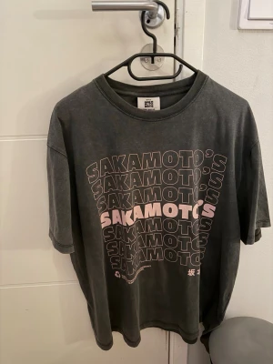 Svart t-shirt med Sakamoto-tryck - Cool svart t-shirt med stort Sakamoto’s-tryck i rosa och vitt på framsidan och japansk butiksgrafik på baksidan. Oversized passform och tillverkad i mjuk bomull. Perfekt för dig som gillar streetwear och unika prints.