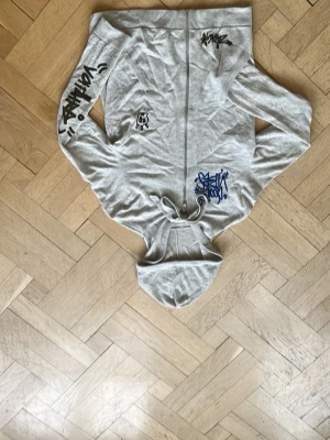 Zadig & Voltare hoodie  - Säljer en grå hoodie med dragkedja och huva. Hoodien har flera coola graffiti-inspirerade tryck i svart, blått och rött på både fram- och baksidan samt på ärmen. Materialet känns mjukt och bekvämt, perfekt för en avslappnad streetwear-look.