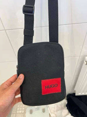Svart axelväska från HUGO - Snygg svart axelväska från HUGO med justerbar rem och röd logga framtill.